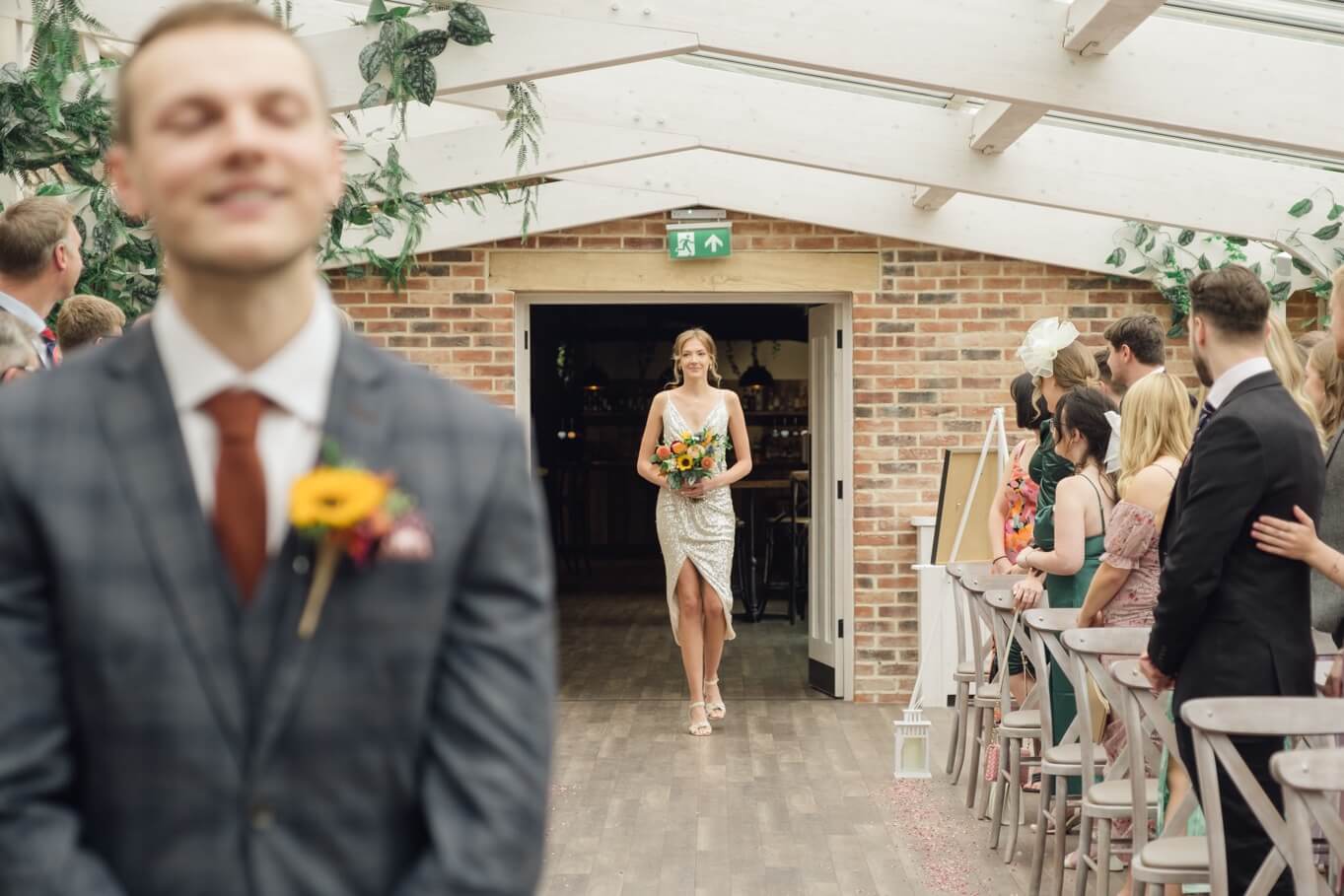 First bridesmaid walking down the aisle in the orangery at Foxtail Barns